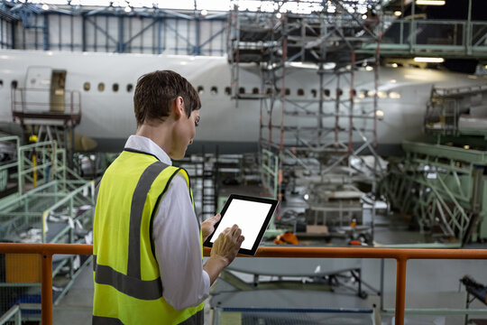Senior female maintenance inspector inspecting jet fuselage on scaffolding in hangar using tablet