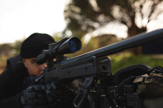 Mature adult male wearing tactical gear lying in park clearing aiming rifle scope mounted on bipod