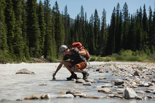 Mid adult male hiker crouching by mountain stream, testing water using container and trekking pole