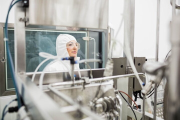 Female technician wearing hooded coverall monitoring steel tubing behind glass partition in lab