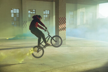Male BMX rider riding black bicycle wheelie emitting yellow green smoke inside abandoned warehouse