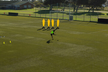 African American male soccer player in green bib kicking ball past mannequins on pitch, copy space
