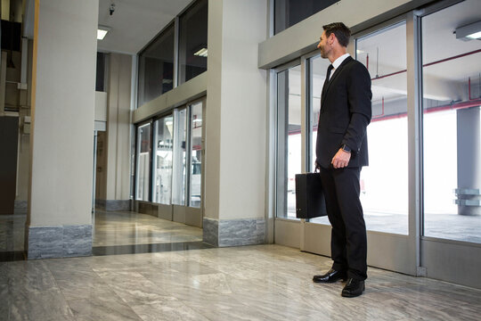 Businessman wearing black suit standing near glass doors in lobby carrying briefcase, copy space