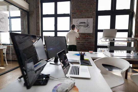 Man studying car illustration pinned on brick wall at design studio with scale model, dual monitors - Powered by Adobe