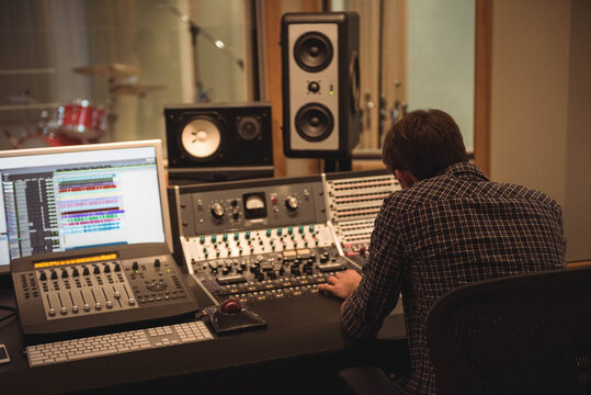 Male sound engineer adjusting faders, knobs on mixing console in studio control room with drum kit