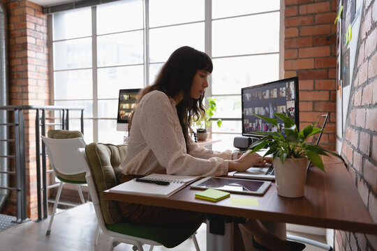 Asian woman sitting at desk in loft office wearing dotted blouse, using laptop and monitor grid