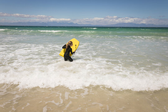 Female child standing knee-deep in foamy surf on sandy beach holding yellow bodyboard facing ocean