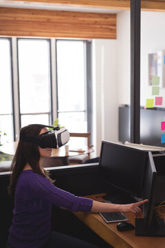 Professional woman wearing VR headset reaching toward dual monitors at modern open-plan office desk