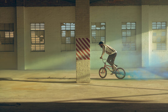 Man riding BMX bike inside warehouse, trailing colored smoke by striped column, copy space - Powered by Adobe