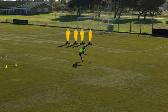 African American male soccer player in green bib kicking ball past mannequins on pitch, copy space