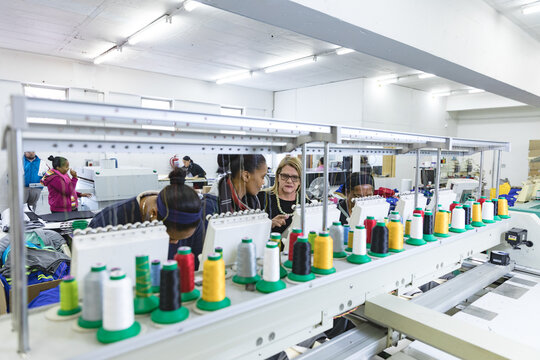 Diverse female staff gathering around embroidery machine instructing and inspecting thread in plant