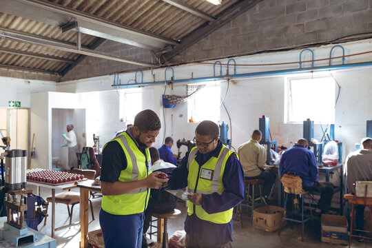 African American male workers inspecting component and noting details on clipboard in workshop