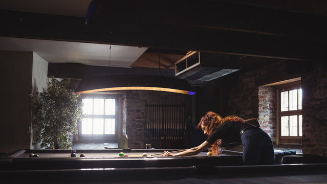 Woman leaning over green pool table aiming cue in game room under lamp, copy space