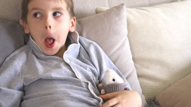 Little European boy of six years old in gray pajamas is sick and coughing. He sits on the sofa and plays with soft toys