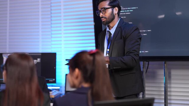 A senior software architect or team lead gives a technical presentation on a new codebase. The screen behind him displays a complex script during a corporate training session.