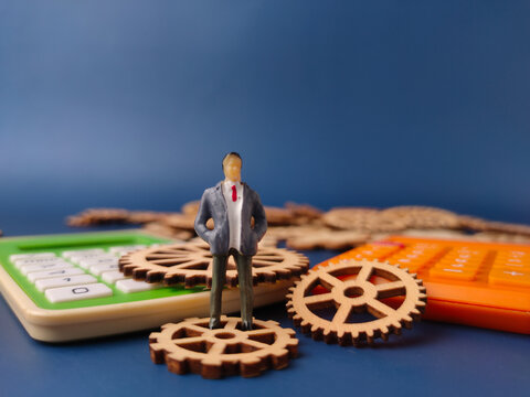 Closeup of a miniature figure of a businessman standing on a wooden gear, with calculators in the foreground