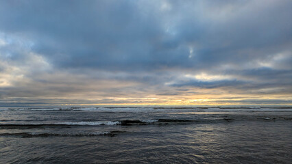 Side view from a wet beach with an emphasis on the small waves rolling in, out westward towards the pacific ocean, under a majestic and complicated cloud-scape near dusk. 