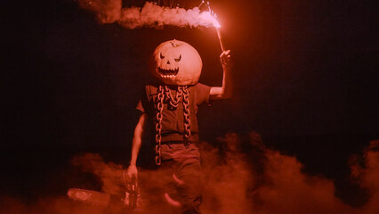 Halloween human pumpkin or Jack o lantern costume on a dark night with lots of smoke, a chainsaw,...