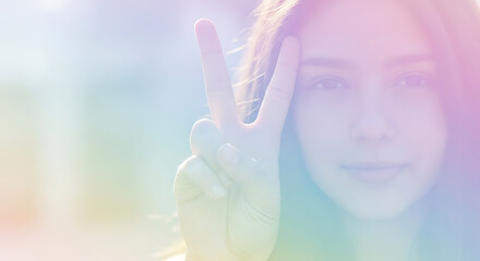 Naklejka premium Young woman with long hair making a peace sign with her fingers, bathed in soft, pastel light.