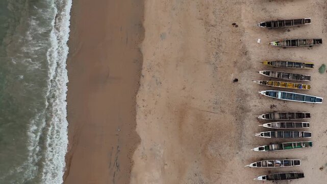 High angle aerial top down shot of Somone fishing beach, Senegal, with wooden boats lined on the shore and waves rolling against the coast.