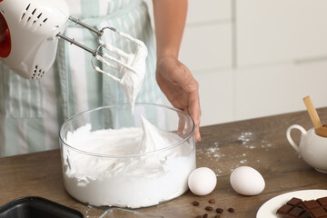 Woman whipping egg albumen with mixer at table in kitchen, closeup