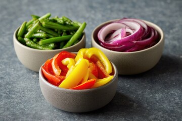 Three small, shallow bowls of chopped vegetables. Green beans, sliced red onion, and sliced bell peppers (yellow & orange) are arranged on a mottled gray surface