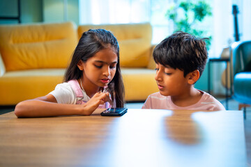 Indian Asian siblings kids using smartphones to play games while enjoying leisure time at home