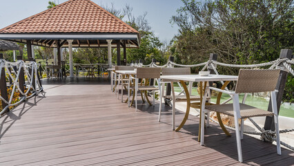 Wicker chairs, tables on the boardwalk terrace outdoors and under a canopy. The pool is visible behind the rope railing. Tropical vegetation. Resort. Hotel. Cuba. Varadero.