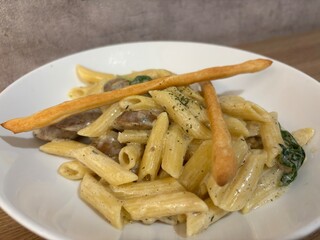 A bowl of creamy penne pasta served in a white dish, topped with sautéed mushrooms and fresh spinach, and garnished with two crispy breadsticks.