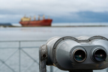 A weathered set of viewing binoculars in sharp focus with a blurred cargo ship in the background across the water under cloudy skies. Nevelsk, Sakhalin Island