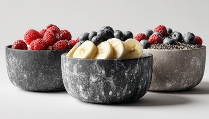 Three bowls of healthy breakfast parfaits.  Each bowl, featuring a speckled, grey-black surface, is filled with a different fruit combination raspberries, blueberries, and bananas.