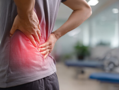Back pain relief exercises demonstrating a person holding their lower back while inside a gym or rehabilitation center with equipment visible in the background.