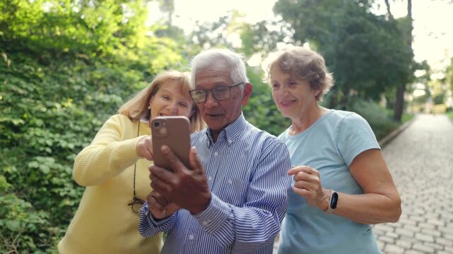 On a sunny day in a lively park, three joyful seniors gather to explore a smartphone, sharing laughs, delightful stories, and cherishing their warm companionship during this special moment