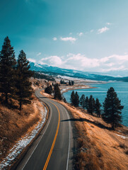 Fototapeta premium Winding road curves along serene lakeside, bordered by tall pine trees and golden grass under clear blue sky. Snow capped mountains rise in distance, creating tranquil landscape