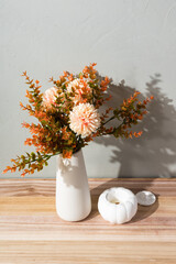 A white vase with autumn flowers stands on a woven placemat, decorated with amber glass candle holders and a white pumpkin. The cozy still life creates a warm seasonal home atmosphere.