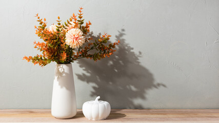 A white vase with autumn flowers stands on a woven placemat, decorated with amber glass candle holders and a white pumpkin. The cozy still life creates a warm seasonal home atmosphere.
