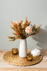 A white vase with autumn flowers stands on a woven placemat, decorated with amber glass candle holders and a white pumpkin. The cozy still life creates a warm seasonal home atmosphere.