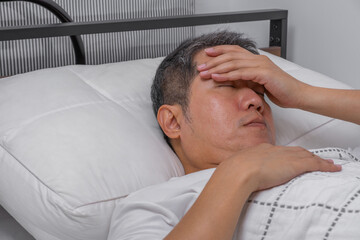 Middle-aged Asian man lying in bed with headache from high blood pressure. Ideal for senior care ads on hypertension treatment, wellness, or medical support.