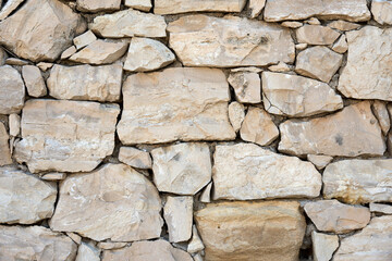 Close-up of a rustic stone wall made of natural beige rocks. Rough texture and irregular shapes create a traditional and sturdy construction background.          