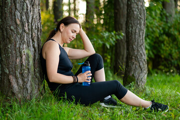 Fototapeta premium Beautiful young woman resting by tree after forest run, holding a water bottle and enjoying nature