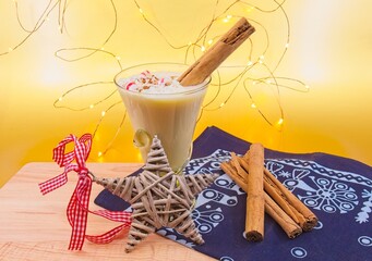Glass of eggnog with whipped cream, cinnamon and peppermint pieces on top. There is a woven star with a red and white ribbon in the foreground. The background is metallic gold and fairy lights.