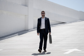 Fototapeta premium Business outdoor portrait. Business man walking outdoors. Successful leader on move. Man in suit with briefcase. Business man going to work. Businessman with briefcase. Business success.