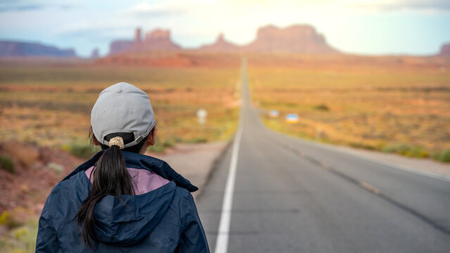 Rear view of Asian female tourist standing off the highway US route 163 looking at the long road view leading to Monument Valley on the Arizona-Utah border, United States. America road trip travel - Powered by Adobe