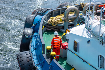 View from the forward section of a large tugboat, featuring a male crew member wearing a red safety...