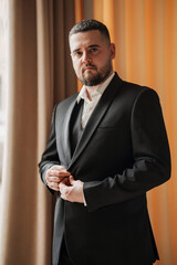 A man with a beard puts on a jacket by the window.