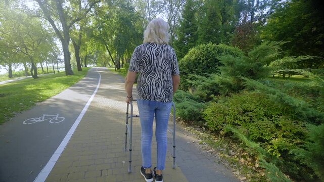 Rear view of senior woman on retirement with age-related mobility disability using walker for walk, helping to maintain balance and stability while enjoying morning stroll in summer park.