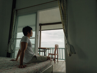 Fototapeta premium Asian male gazing through open window at cloudy morning sky from cozy bedroom interior