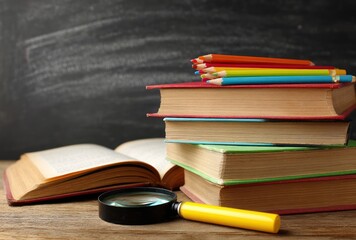 Stacked books and colored pencils are arranged beside an open book with a magnifying glass, sitting atop a wooden surface, and a chalkboard backdrop