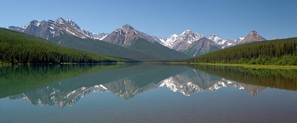 Tranquil lake reflects snow-capped mountains under a clear blue sky, framed by lush green forests on either side, creating a serene and mirrored landscape