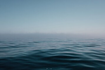 Fototapeta premium Calm sea ripples under a pale blue sky partially veiled in mist, evoking tranquility and vastness. Low horizon line